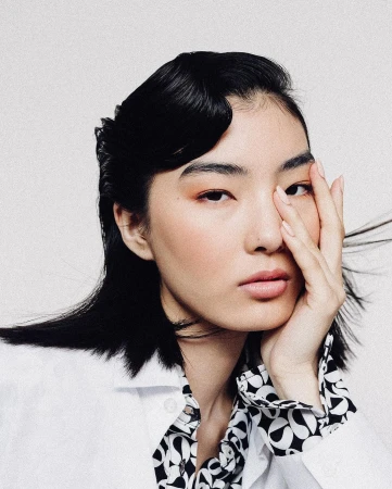 Portrait of a woman with black hair and hand near face wearing a patterned shirt against a white backdrop