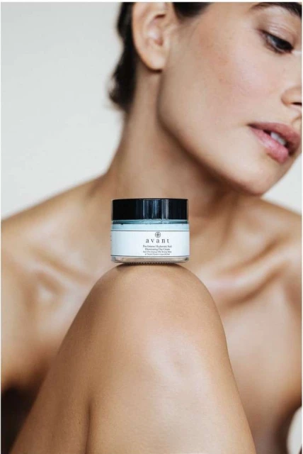 Close-up shot of a light-skinned female with a skin cream jar resting on her knee, set against a light background in a beauty context.