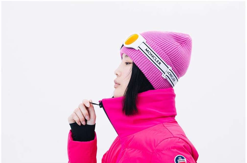 Close-up of an Asian female model wearing a pink Moncler beanie and matching puffer jacket against a bright white background.