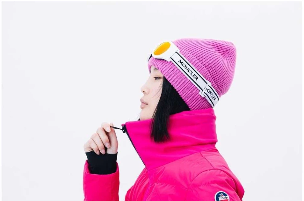 Close-up of an Asian female model wearing a pink Moncler beanie and matching puffer jacket against a bright white background.