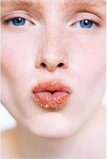 Close-up portrait of a fair-skinned adult woman with blue eyes and freckles, wearing sugar scrub on her lips, set against a bright background.