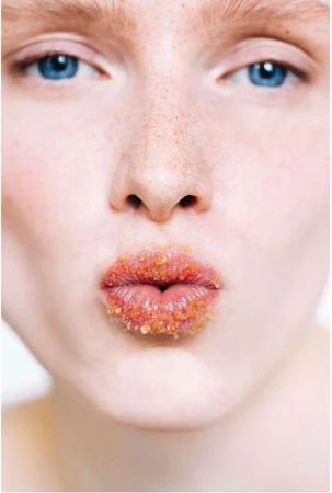 Close-up portrait of a fair-skinned adult woman with blue eyes and freckles, wearing sugar scrub on her lips, set against a bright background.