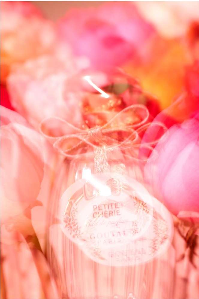 Abstract long exposure photography of 'Petite Cherie' perfume bottle surrounded by blurred pink flowers creates a dreamy, ethereal effect.