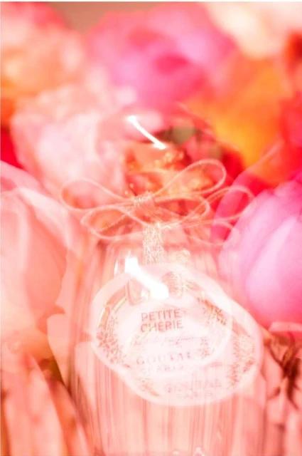 Abstract long exposure photography of 'Petite Cherie' perfume bottle surrounded by blurred pink flowers creates a dreamy, ethereal effect.