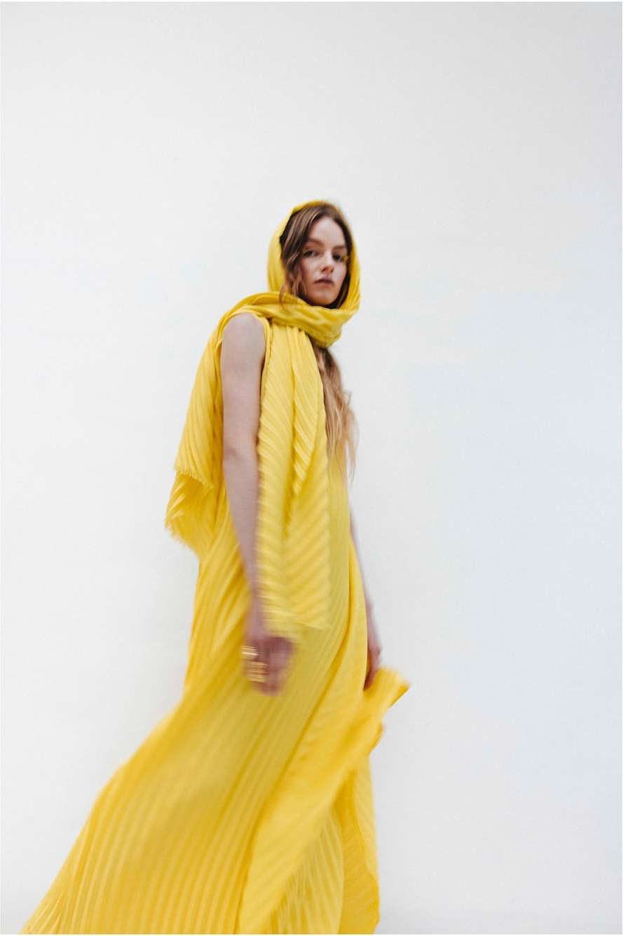 Woman in a yellow pleated dress and headscarf poses in front of a white backdrop in a fashion or editorial style photograph.