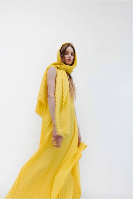 Woman in a yellow pleated dress and headscarf poses in front of a white backdrop in a fashion or editorial style photograph.