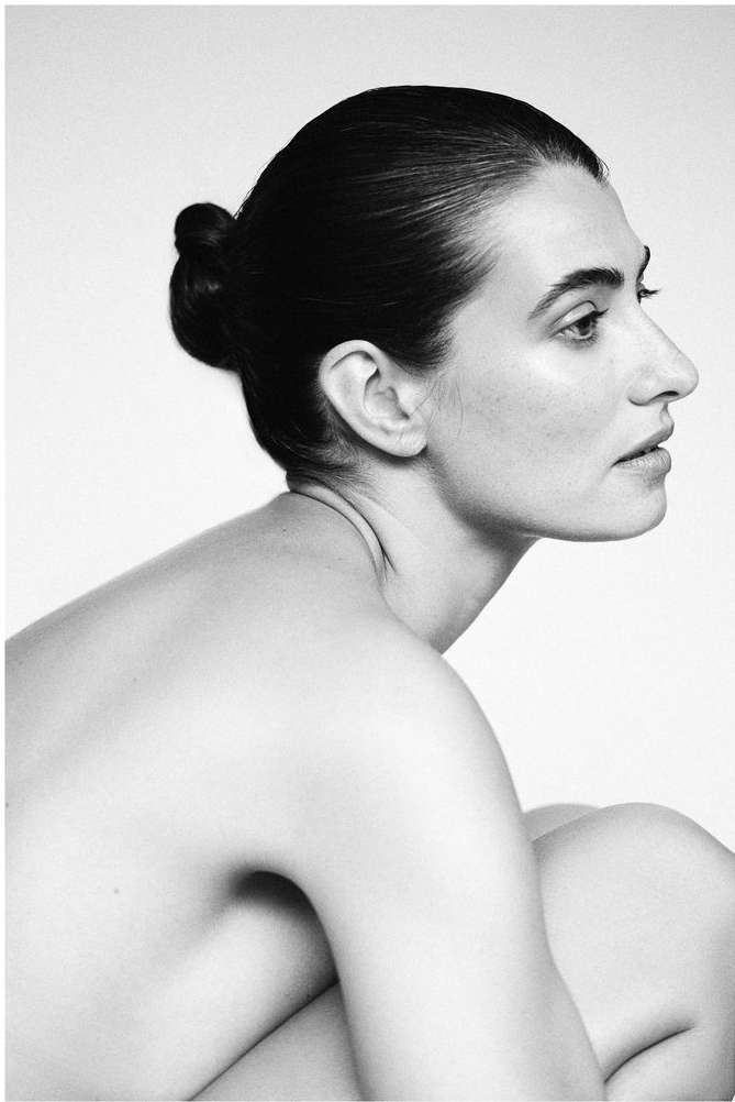 Monochrome portrait of a woman with dark hair in a bun, looking to the side against a bright background in a photo studio setting.