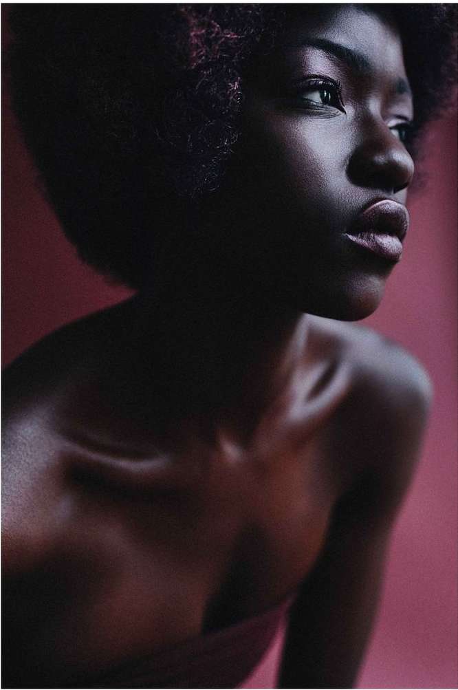 Portrait of a young, dark-skinned woman with curly hair against a light red background. She is wearing makeup and looking off to the side.