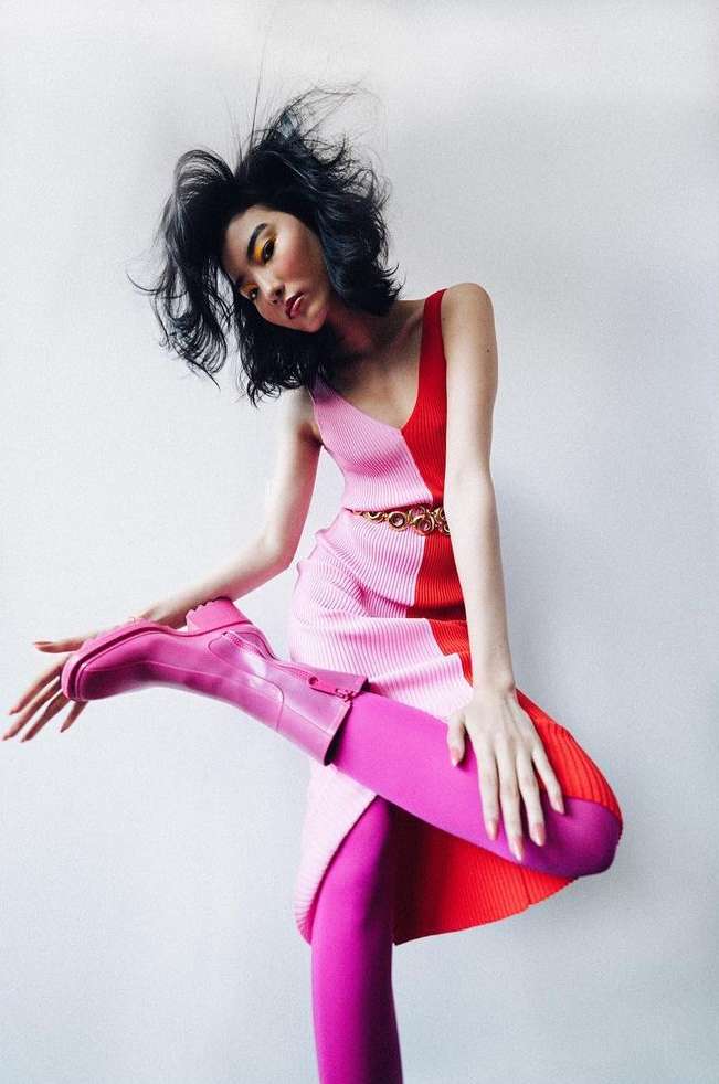 Woman in pink-and-red color-blocked dress and pink tights and boots striking a fashion pose in front of a white wall.