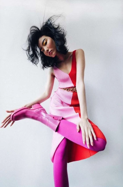 Woman in pink-and-red color-blocked dress and pink tights and boots striking a fashion pose in front of a white wall.