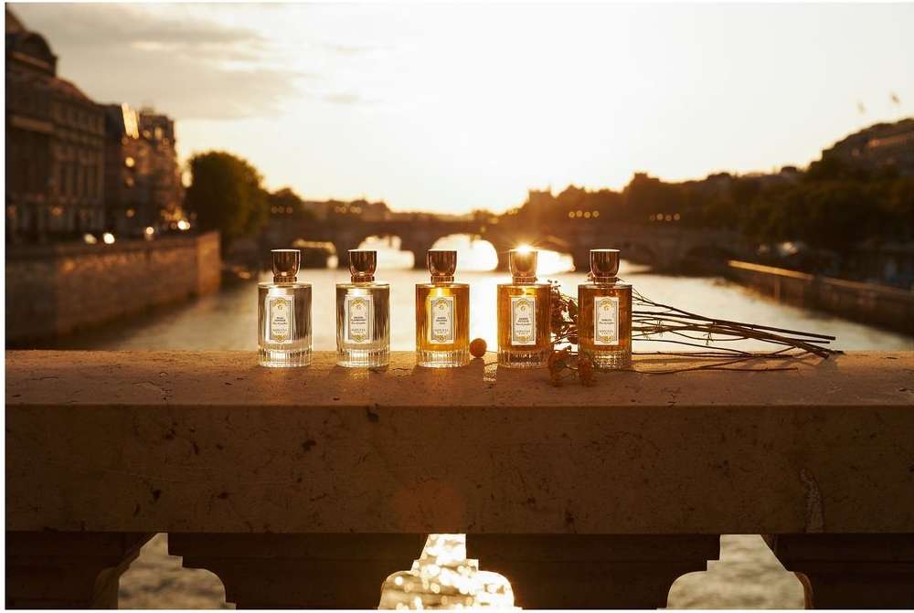 Five perfume bottles are lined up on a ledge with a river and city buildings in the background during a golden sunset.