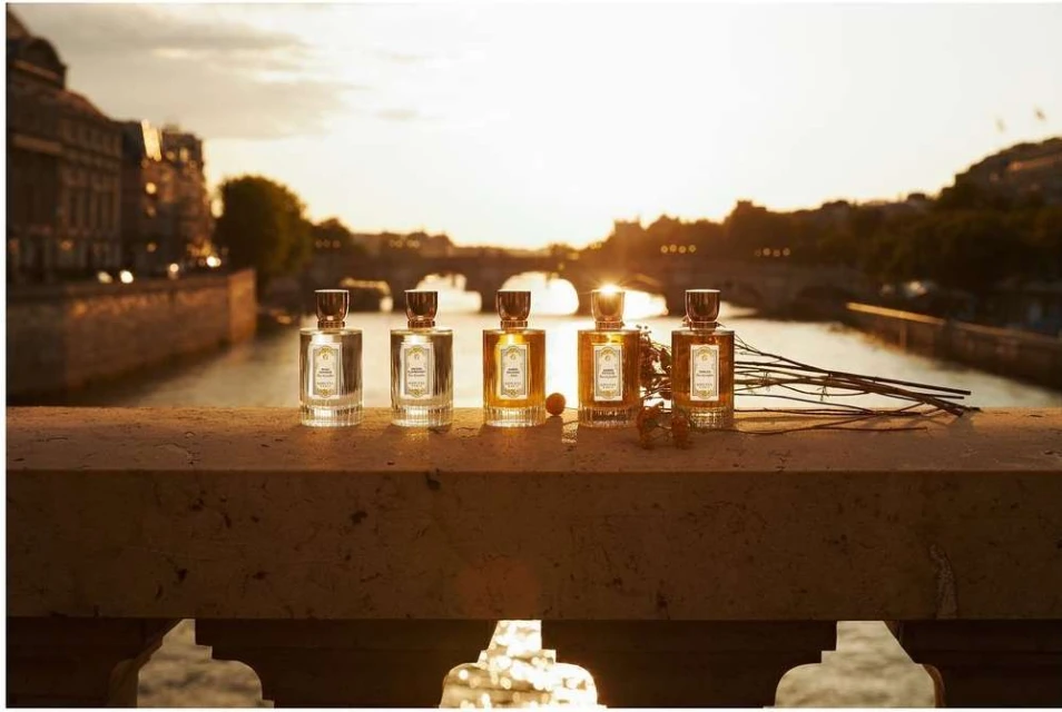 Five perfume bottles are lined up on a ledge with a river and city buildings in the background during a golden sunset.