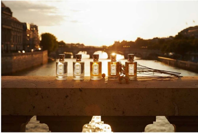 Five perfume bottles are lined up on a ledge with a river and city buildings in the background during a golden sunset.