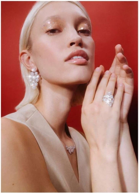Portrait of a blonde woman with glittery eyeshadow, wearing earrings, ring, and necklace with gemstones against a red background.