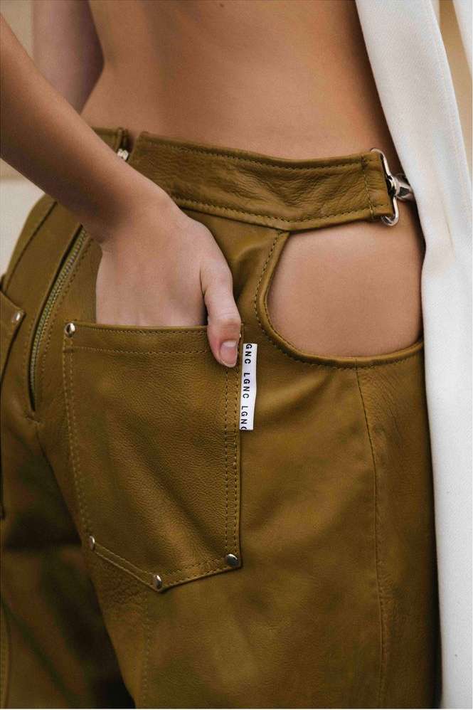Close-up of an adult with their hand in the pocket of brown, high-waisted leather pants with an open back and white fabric detail.