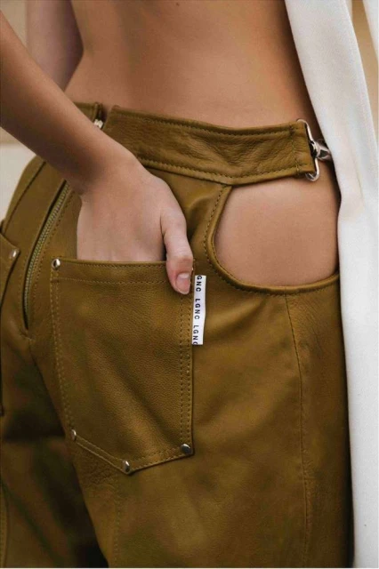 Close-up of an adult with their hand in the pocket of brown, high-waisted leather pants with an open back and white fabric detail.