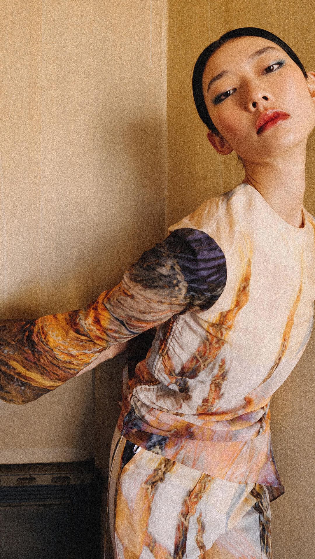 Portrait of an Asian woman with a neutral expression wearing a patterned top and skirt, posing indoors with natural lighting.