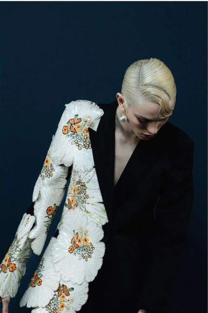 Headshot of a blonde woman wearing a black blazer with ornate floral appliqués, set against a dark blue backdrop in a fashion editorial.