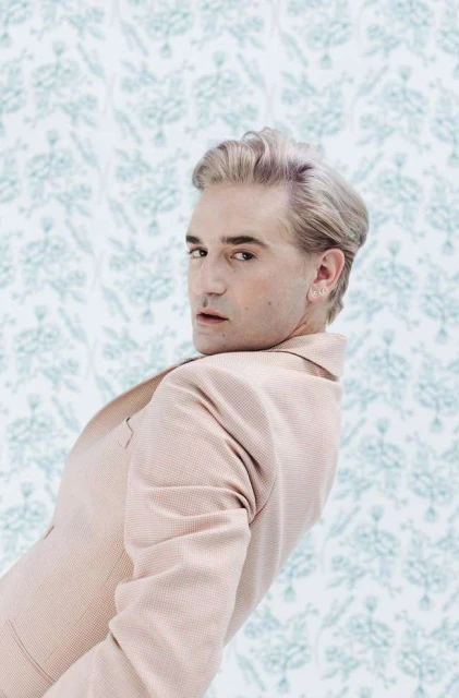 Stylish adult male with silver hair poses in a peach suit against a blue patterned backdrop in a studio setting, looking over shoulder.