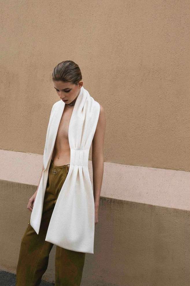 A fashionable young woman in a draped white outfit poses outdoors against a neutral wall, looking down with a shallow depth of field.