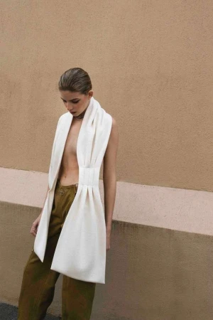 A fashionable young woman in a draped white outfit poses outdoors against a neutral wall, looking down with a shallow depth of field.
