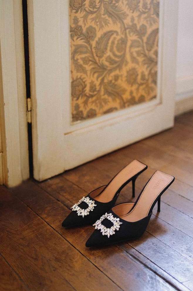 Elegant black mule heels with sparkling embellishments displayed on a wooden floor near a vintage door showcasing patterned wallpaper.