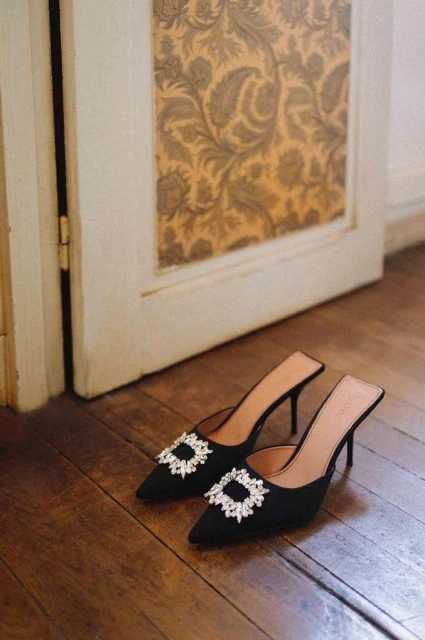 Elegant black mule heels with sparkling embellishments displayed on a wooden floor near a vintage door showcasing patterned wallpaper.