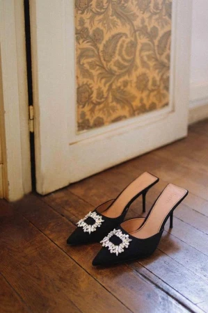 Elegant black mule heels with sparkling embellishments displayed on a wooden floor near a vintage door showcasing patterned wallpaper.