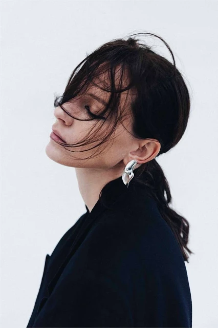 Profile shot of a woman with dark hair wearing silver earrings and a black jacket against a bright white background in a photo studio.