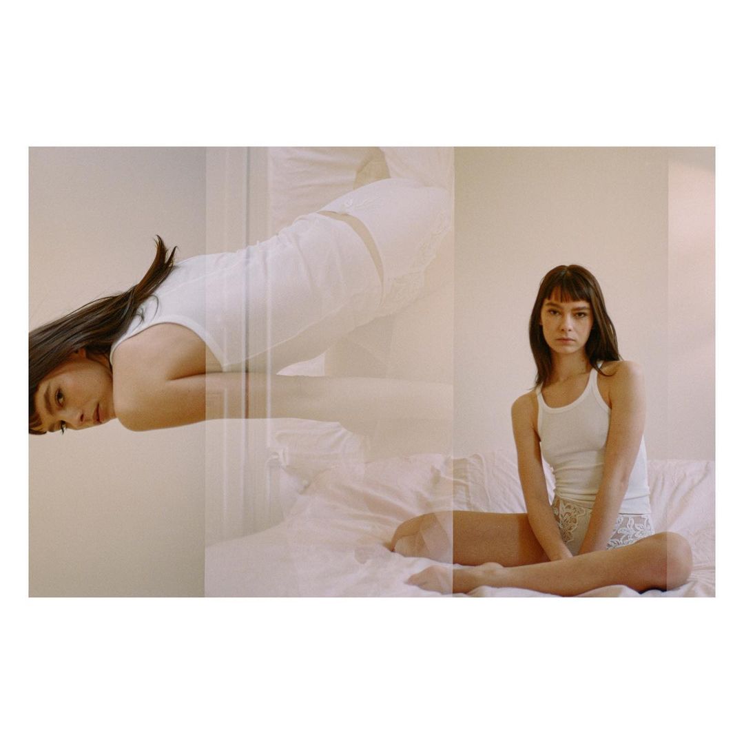 An adult female with dark hair poses sitting in bed in a white tank top with a double exposure, creating a dreamy, intimate vibe.