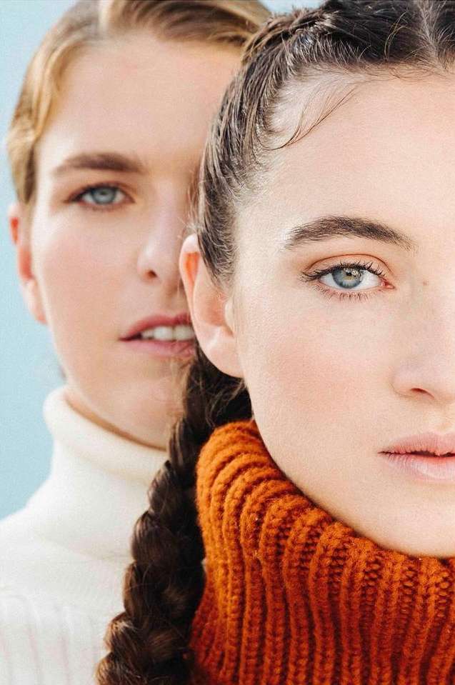 Two female fashion models pose indoors, one in a cream turtleneck and the other in an orange turtleneck, against a light blue background.