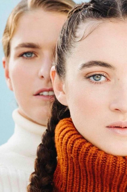 Two female fashion models pose indoors, one in a cream turtleneck and the other in an orange turtleneck, against a light blue background.