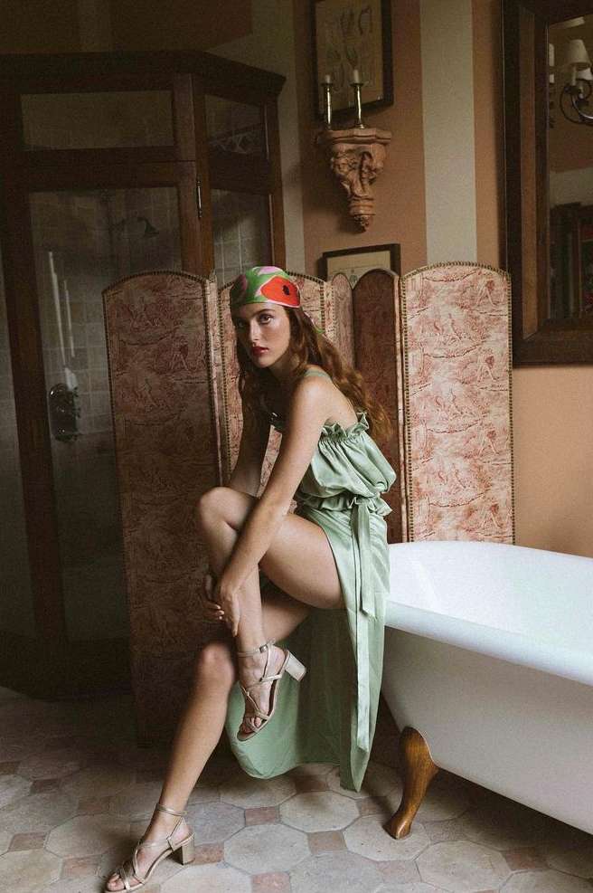 A fair-skinned woman in a green dress and patterned headscarf sits on the edge of a vintage bathtub in an ornate bathroom setting.
