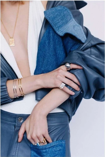 A person wears a blue leather jacket, silver rings, and gold jewelry while posing with a hand in the denim pocket of blue pants.