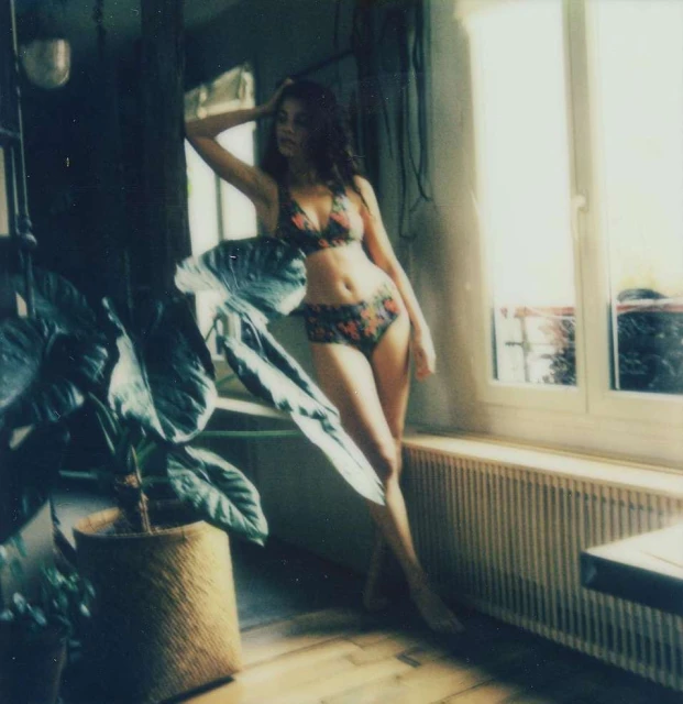 A woman in a floral bikini poses near a window with a large potted plant, creating a bright and natural indoor fashion shot.