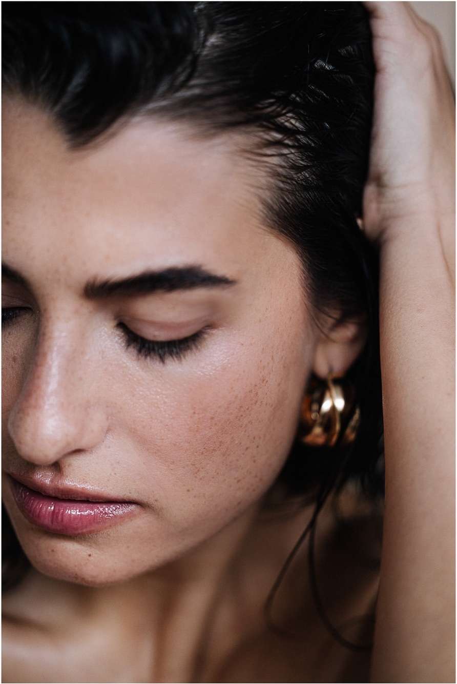 Close-up portrait of an adult woman with dark hair and gold hoop earrings, looking down with a serene expression. Beauty shot, indoor setting.