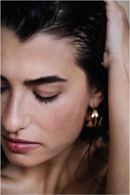 Close-up portrait of an adult woman with dark hair and gold hoop earrings, looking down with a serene expression. Beauty shot, indoor setting.