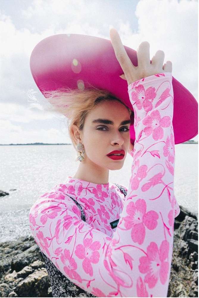 Woman in floral top and oversized hat poses outdoors near rocky beach under partly cloudy sky, wearing statement earrings and red lipstick.