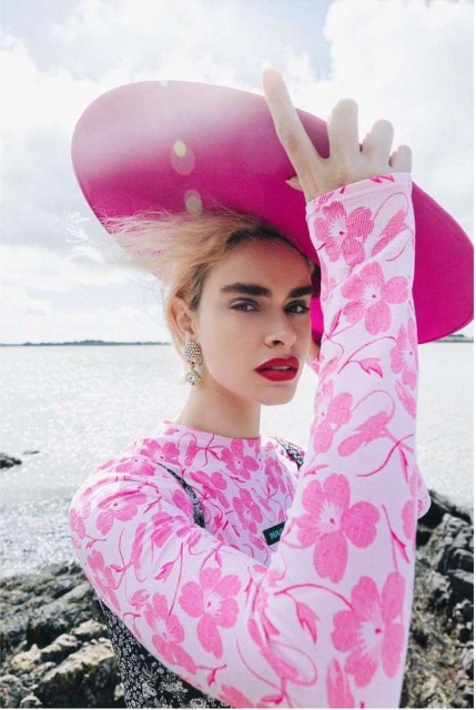 Woman in floral top and oversized hat poses outdoors near rocky beach under partly cloudy sky, wearing statement earrings and red lipstick.
