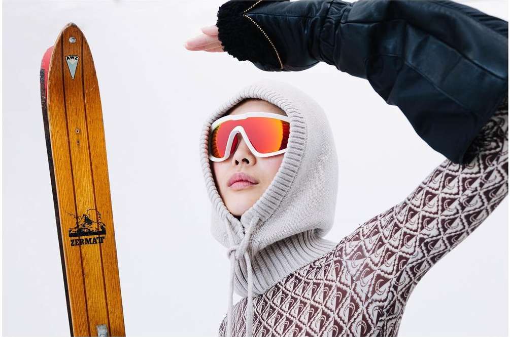 Woman in a knit hood and red goggles poses with a vintage wood ski. Bright indoor fashion shot with a snowy winter sports vibe.