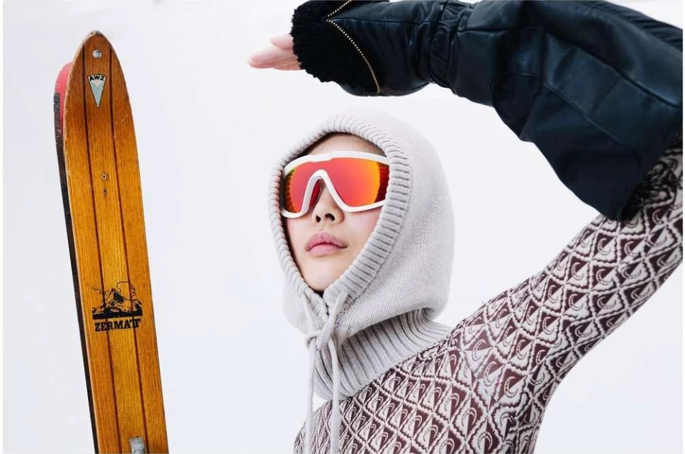 Woman in a knit hood and red goggles poses with a vintage wood ski. Bright indoor fashion shot with a snowy winter sports vibe.