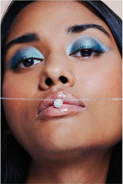 Close-up portrait of an adult woman with dramatic blue eyeshadow wearing a silver necklace with a flower-shaped pendant on her lip.