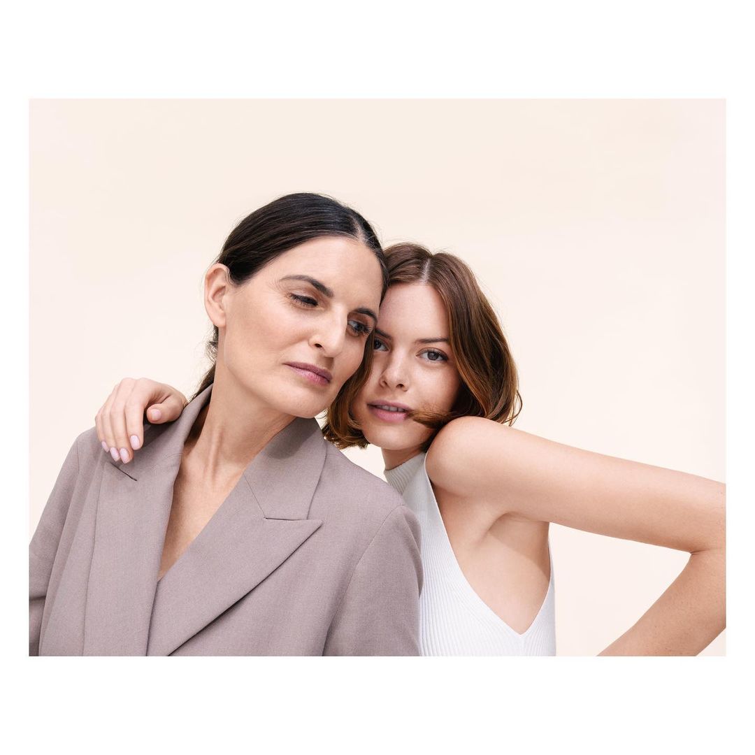 Two women in a portrait embracing. The woman in front wears a white top. The woman behind wears a grey blazer. Neutral background.