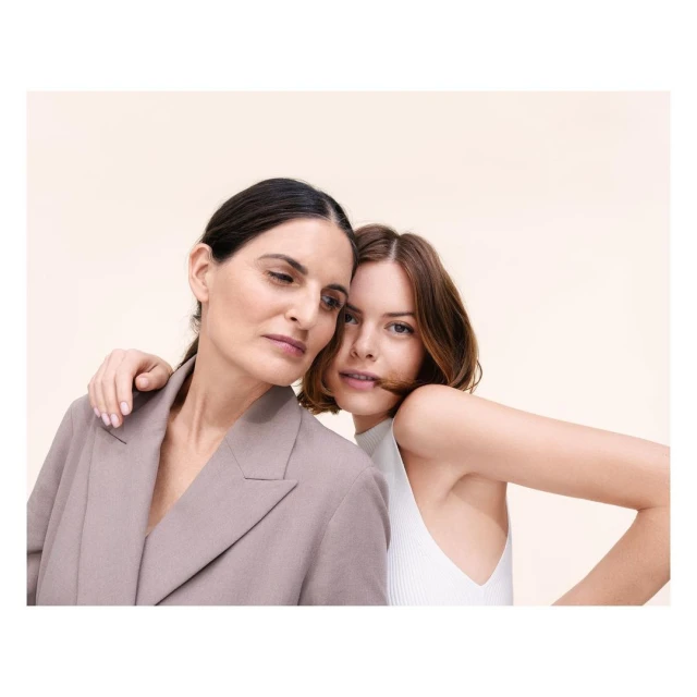 Two women in a portrait embracing. The woman in front wears a white top. The woman behind wears a grey blazer. Neutral background.