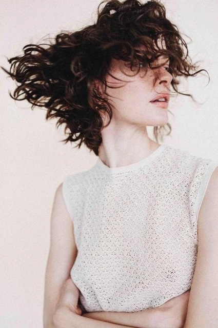Portrait of a stylish brunette woman with curly hair blowing. She is wearing a cream-colored top and posing in a photo studio.