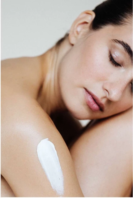 Close-up of a bare-shouldered woman with her eyes closed, a patch of lotion on her arm against a simple, neutral background.