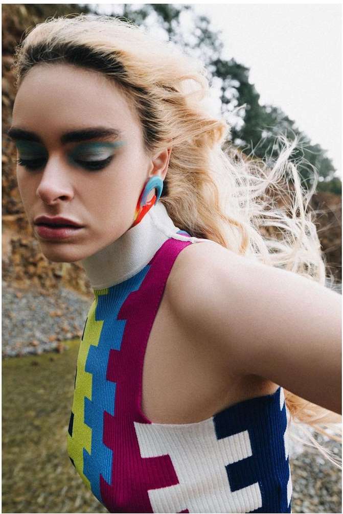 A fair-skinned woman with blonde hair poses in a colorful top outdoors. The photo features unique makeup and earrings, captured at eye level.