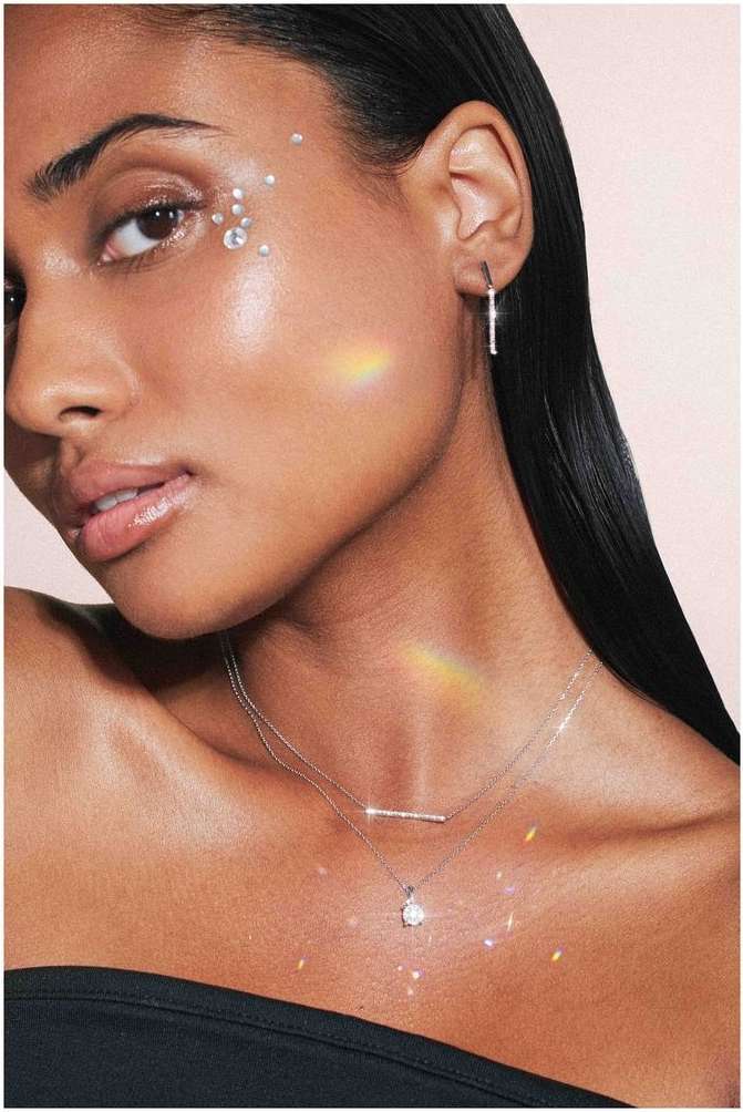 Close-up of a woman with face gems, wearing jewelry and a black strapless top against a light pink background in an indoor setting.