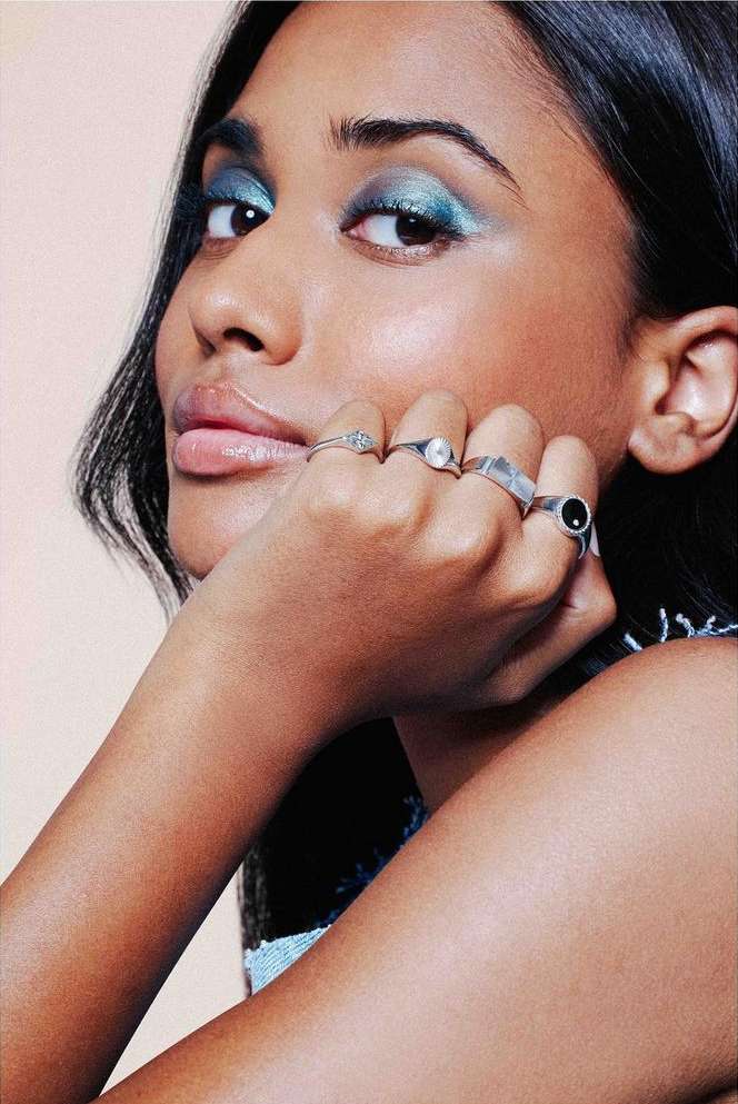 Close-up portrait of a woman with teal eyeshadow and silver rings on her fingers posing against a light tan backdrop.