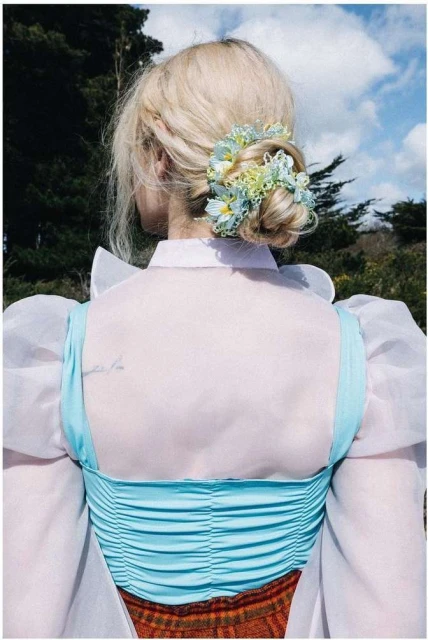 Blonde woman with updo hairstyle, adorned with light blue flowers, wears light blue top and sheer white sleeves, against a backdrop of trees.